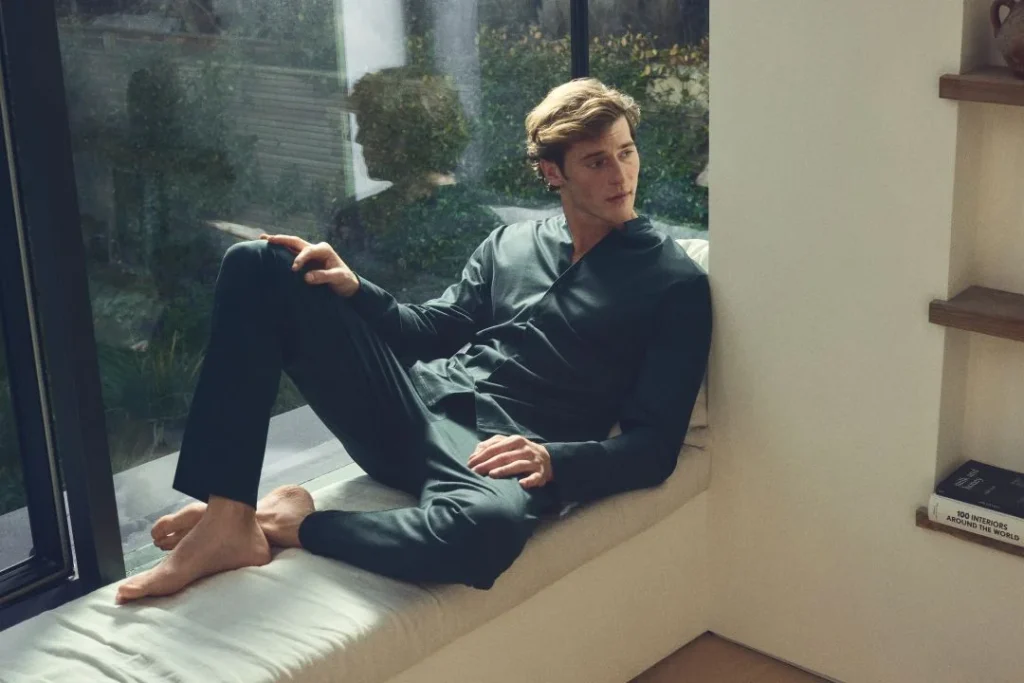 Homem de pijama escuro sentado descalço na janela.