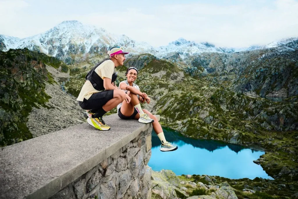 Dois corredores de trilho descansam perante montanhas e lago azul.