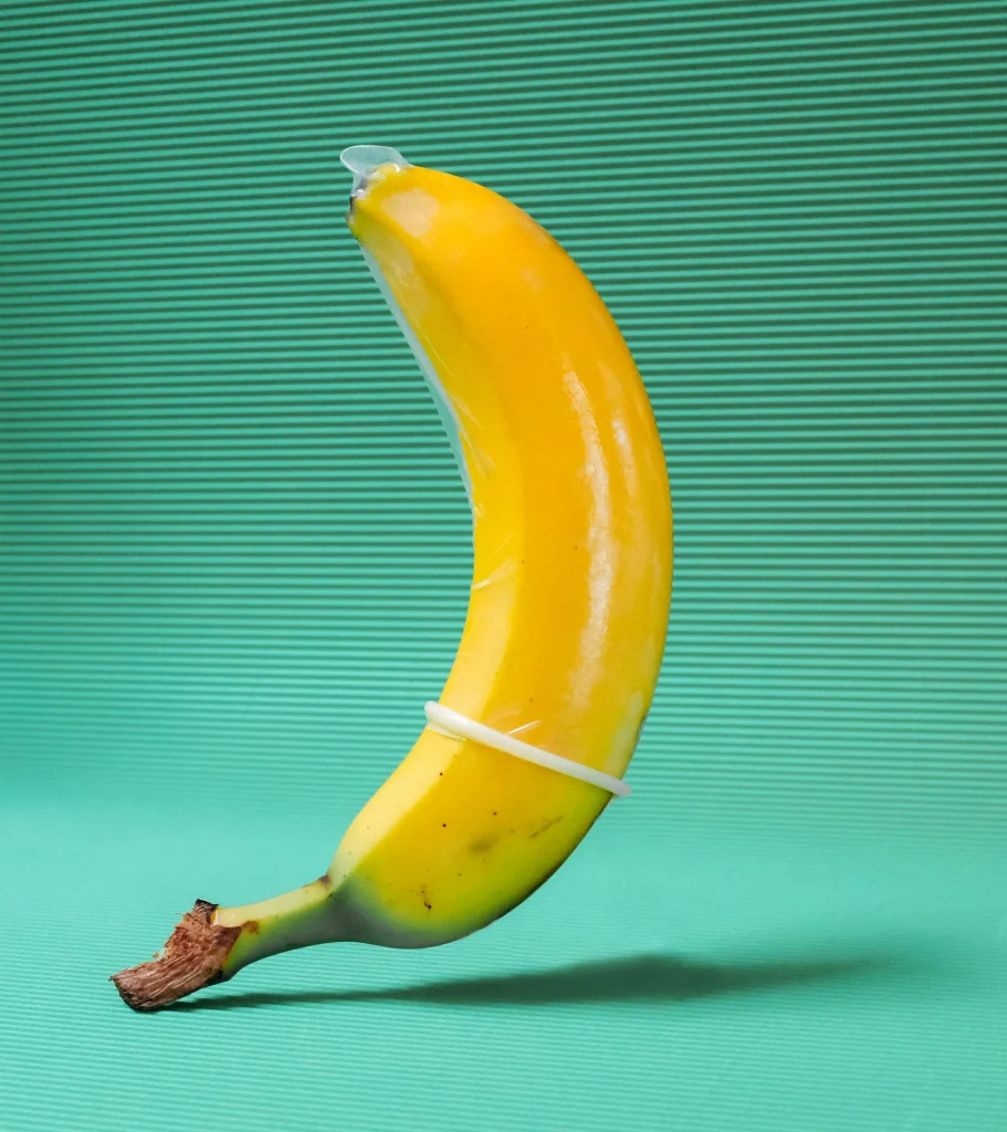 Banana amarela com um preservativo desenrolado até meio sobre um fundo verde texturizado, representando a largura nominal.