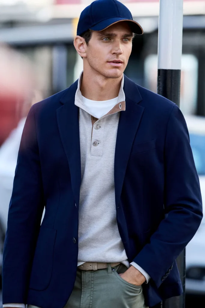 Homem veste blazer azul marinho, polo bege e boné da coleção Hackett London outono inverno
