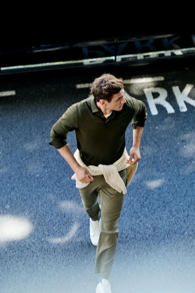 Homem a caminhar numa rua asfaltada a usar um polo de manga comprida verde escuro, calças verdes e uma camisola clara atada à cintura da coleção Hackett London outono inverno