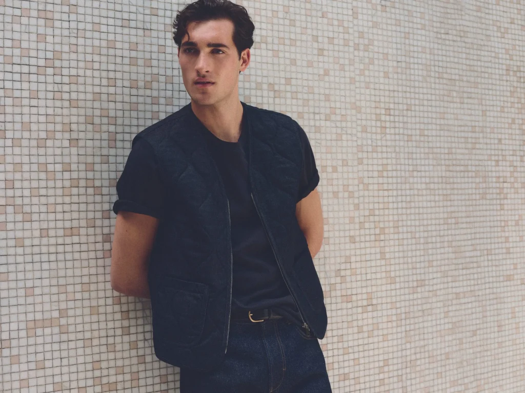 Modelo masculino encostado a uma parede a vestir colete acolchoado escuro, t-shirt e calças da coleção Mango Man Superior Denim