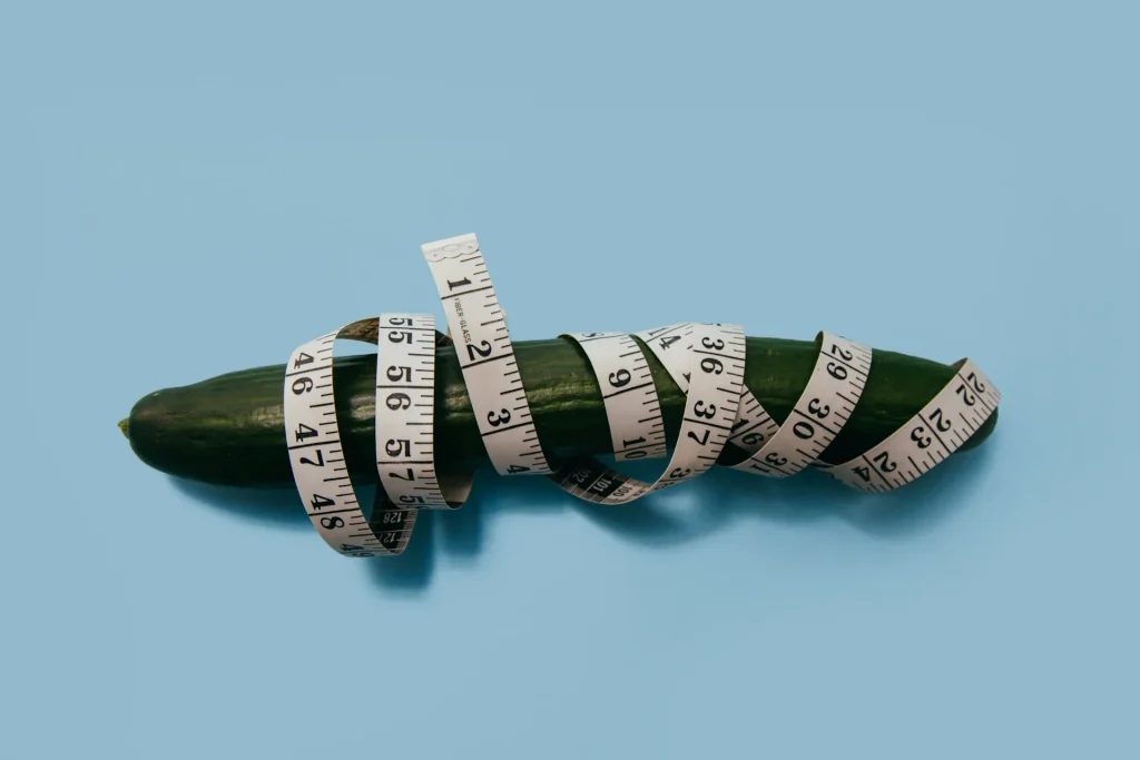Pepino verde enrolado numa fita métrica branca sobre fundo azul claro ilustrando como medir a grossura do pénis
