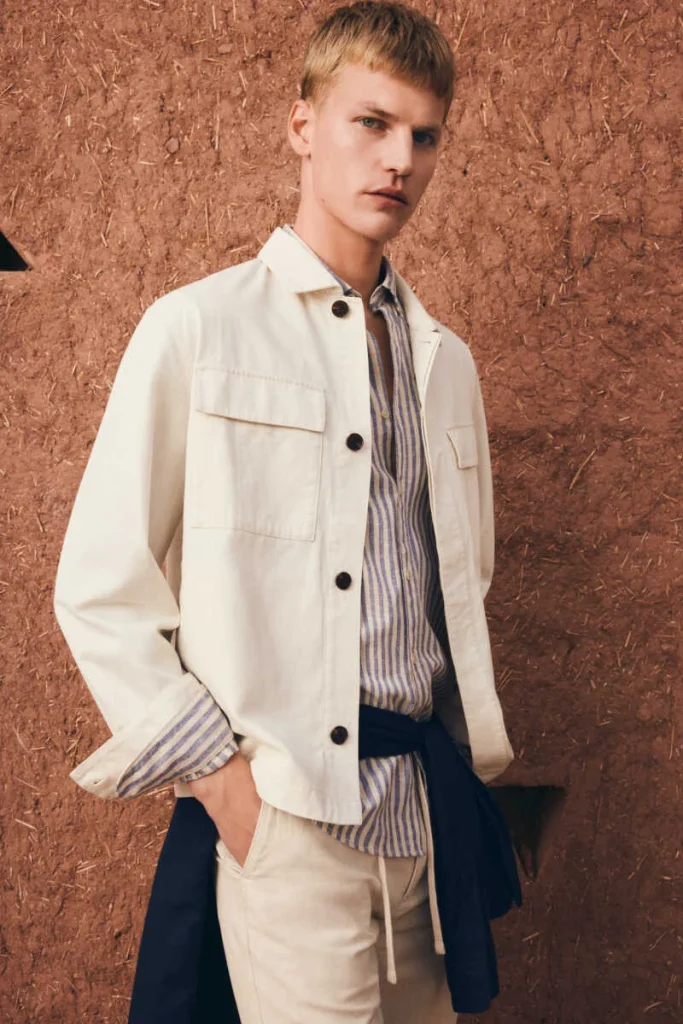 Homem com casaco creme e camisa às riscas em parede rústica.