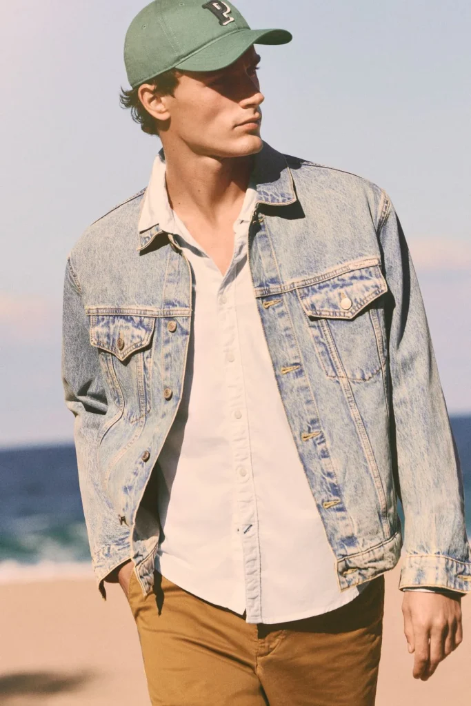 Homem na praia com casaco ganga, camisa branca e boné verde.