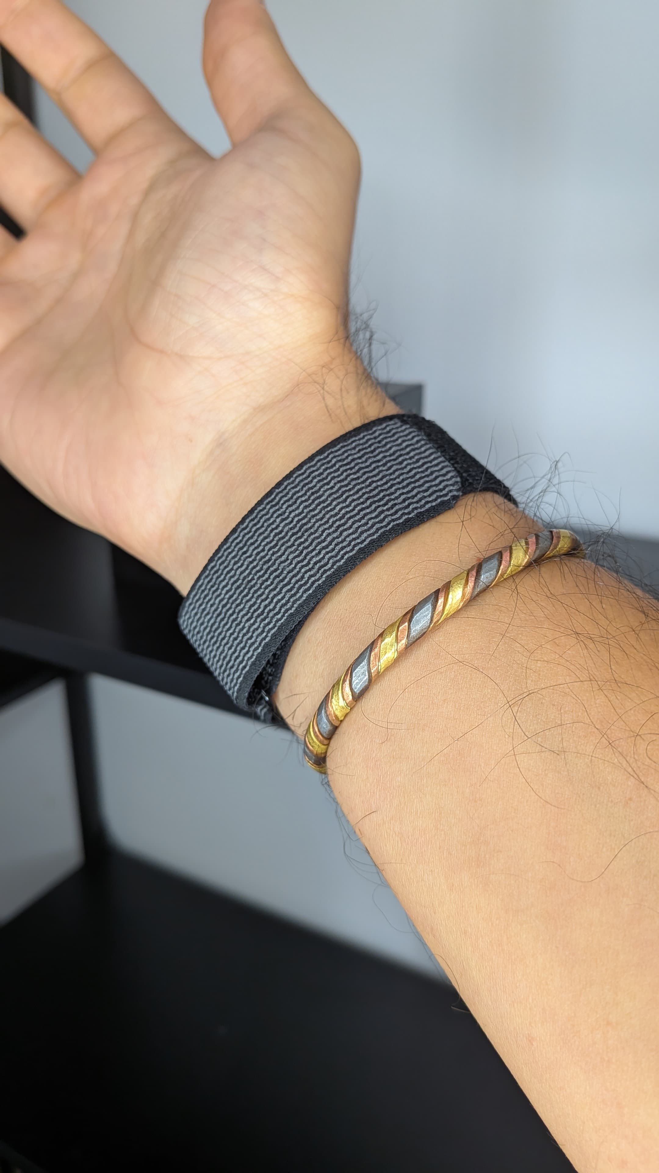 Pulso de um homem com a pulseira desportiva duckdik sense de tecido preto sem ecrã ao lado de uma pulseira metálica dourada e prateada.