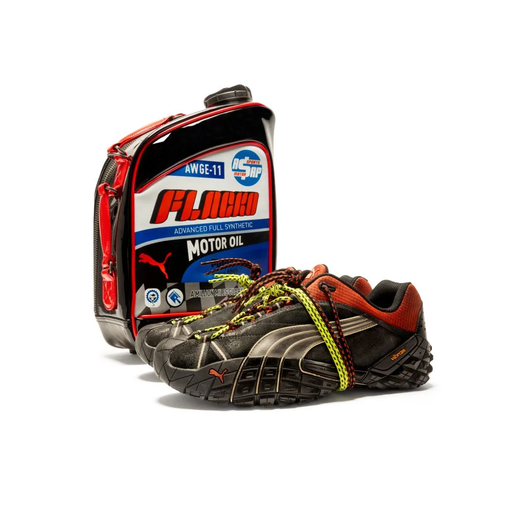 Sapatilhas pretas e vermelhas Puma Straycat A$AP Rocky com atacadores duplos e sola Vibram junto a uma mala em forma de lata de óleo de motor