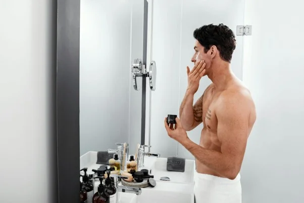 Homem em tronco nu em frente ao espelho da casa de banho a aplicar creme no rosto durante uma rotina de skincare coreana.