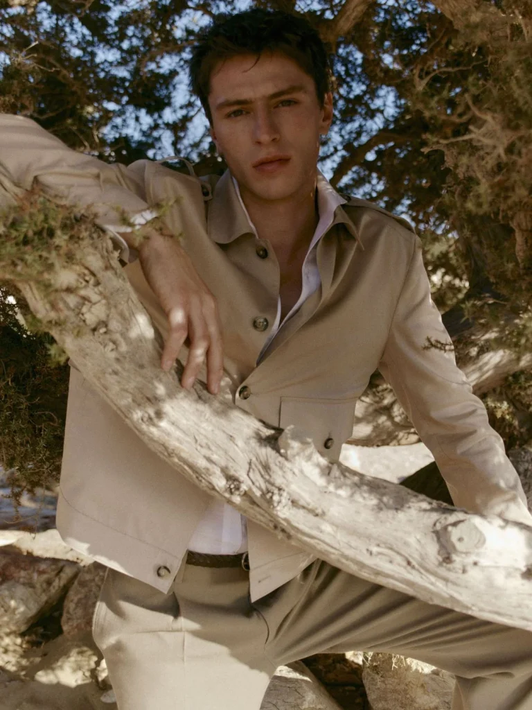 Homem de casaco bege a posar em tronco de árvore.