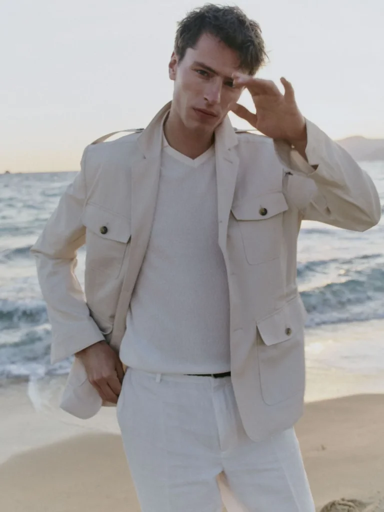 Homem elegante na praia, casaco bege, calças brancas, sol.