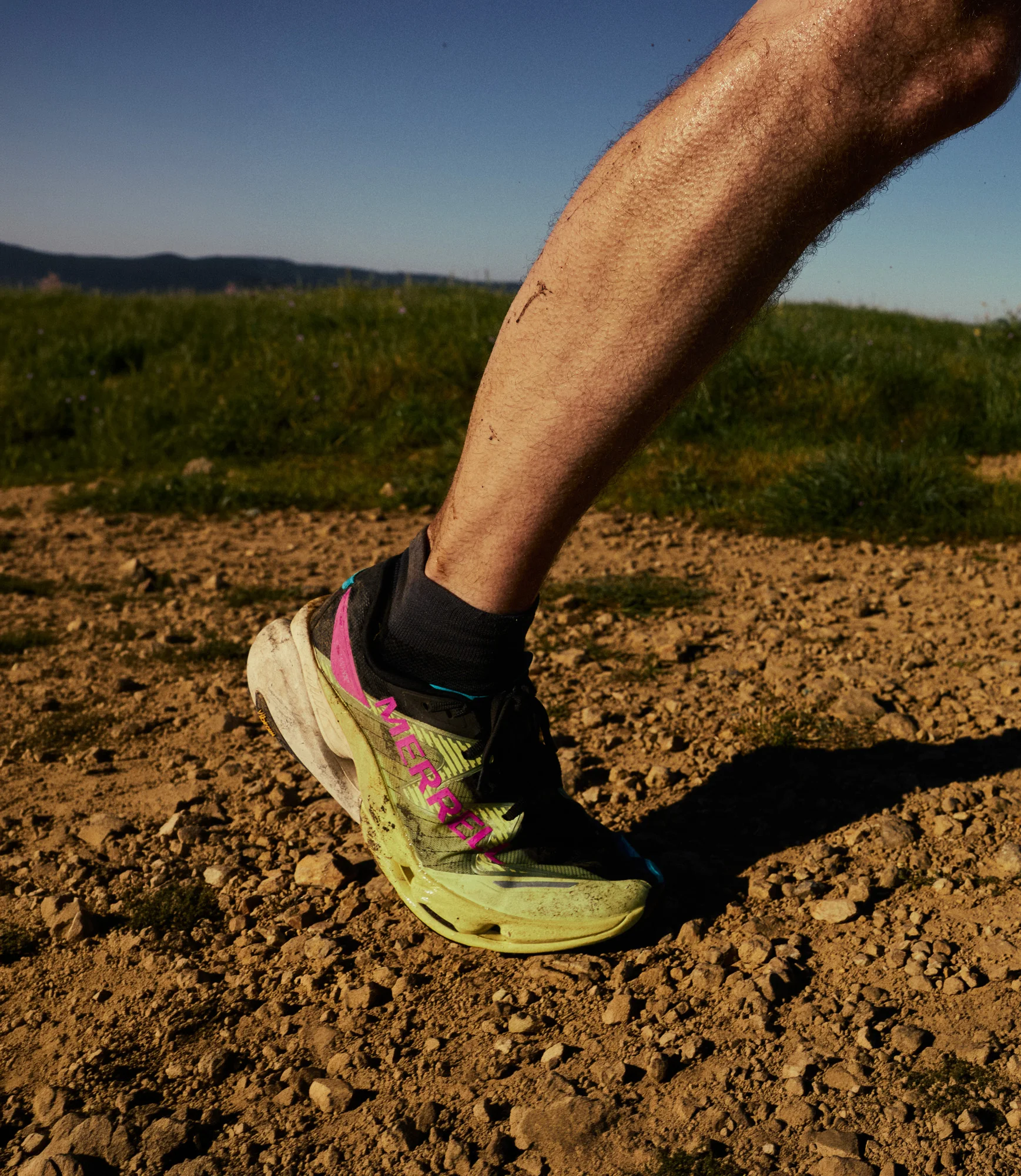 Pé de um homem a pisar um trilho de terra e pedras com as sapatilhas de trail Merrell MTL SpeedARC Peak em tons de amarelo, preto e rosa