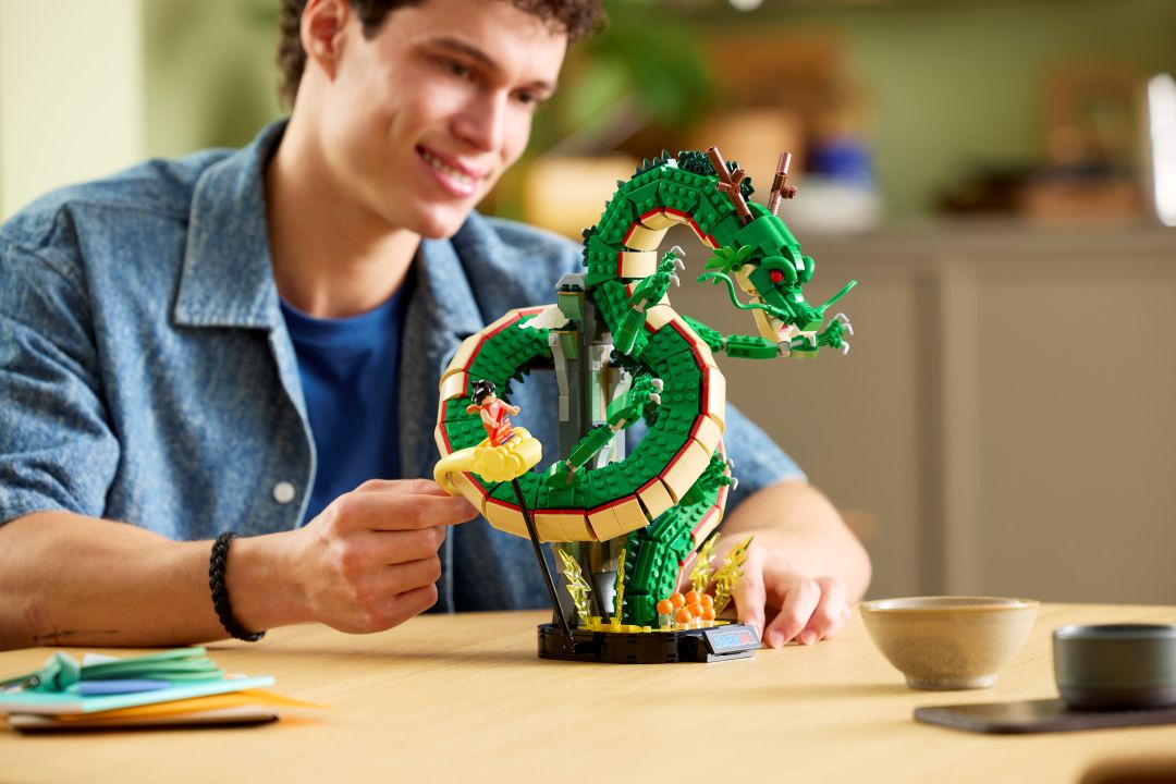 Jovem a interagir com o set LEGO Dragon Ball montado sobre uma mesa de madeira, destacando o corpo verde do dragão Shenron e a minifigura do Songoku