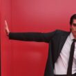 Homem de fato preto contra parede vermelha, pose elegante.