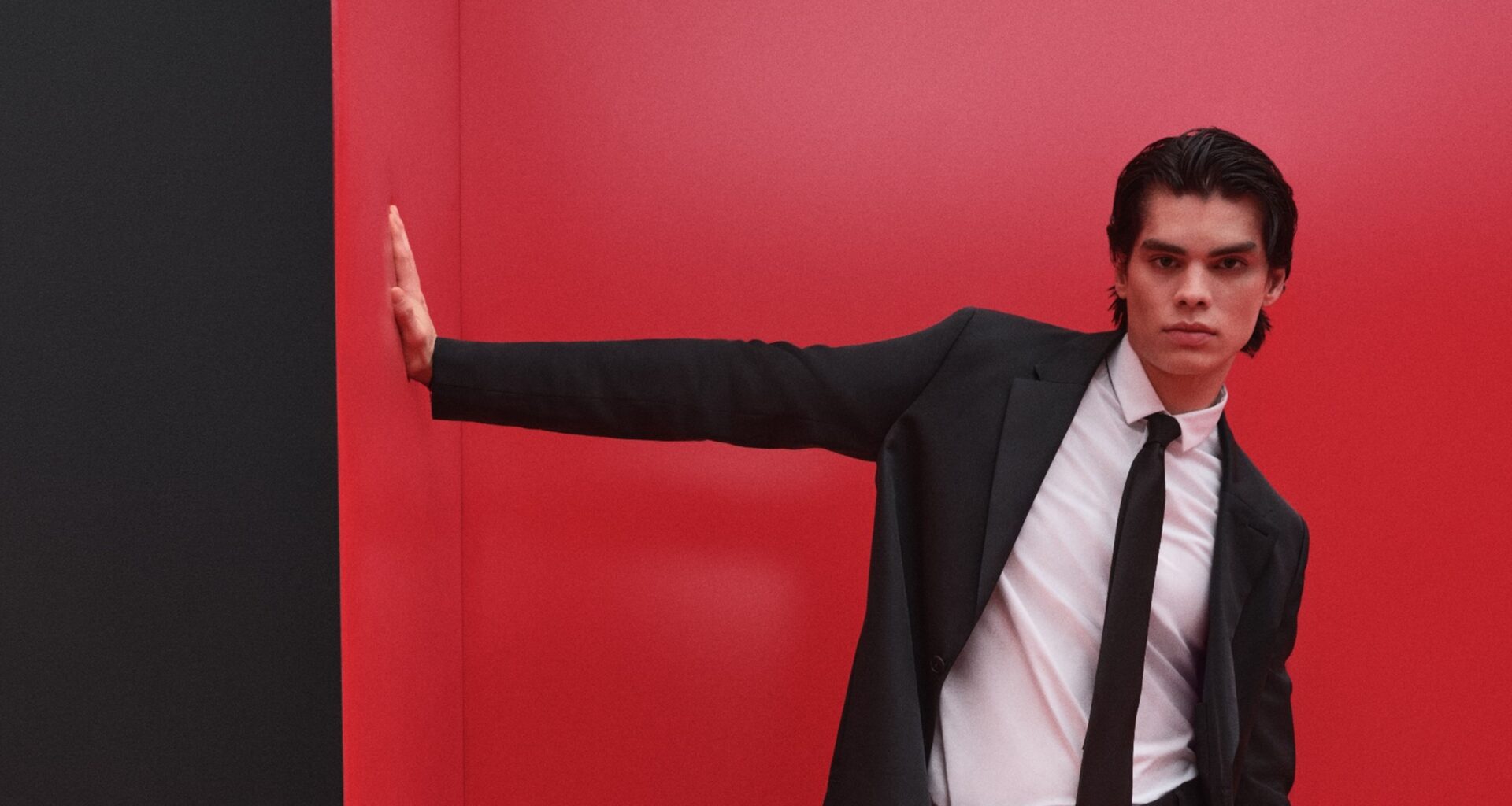 Homem de fato preto contra parede vermelha, pose elegante.