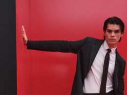 Homem de fato preto contra parede vermelha, pose elegante.