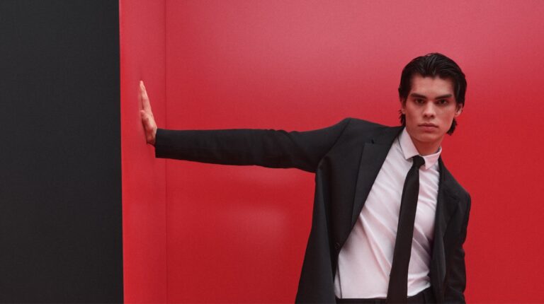 Homem de fato preto contra parede vermelha, pose elegante.