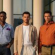Três homens com estilo a posar num ambiente urbano, vestindo as principais tendências de moda masculina 2026, incluindo alfaiataria desconstruída, padrões Gingham e Zebra, e uma paleta de cores solares.