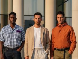 Três homens com estilo a posar num ambiente urbano, vestindo as principais tendências de moda masculina 2026, incluindo alfaiataria desconstruída, padrões Gingham e Zebra, e uma paleta de cores solares.