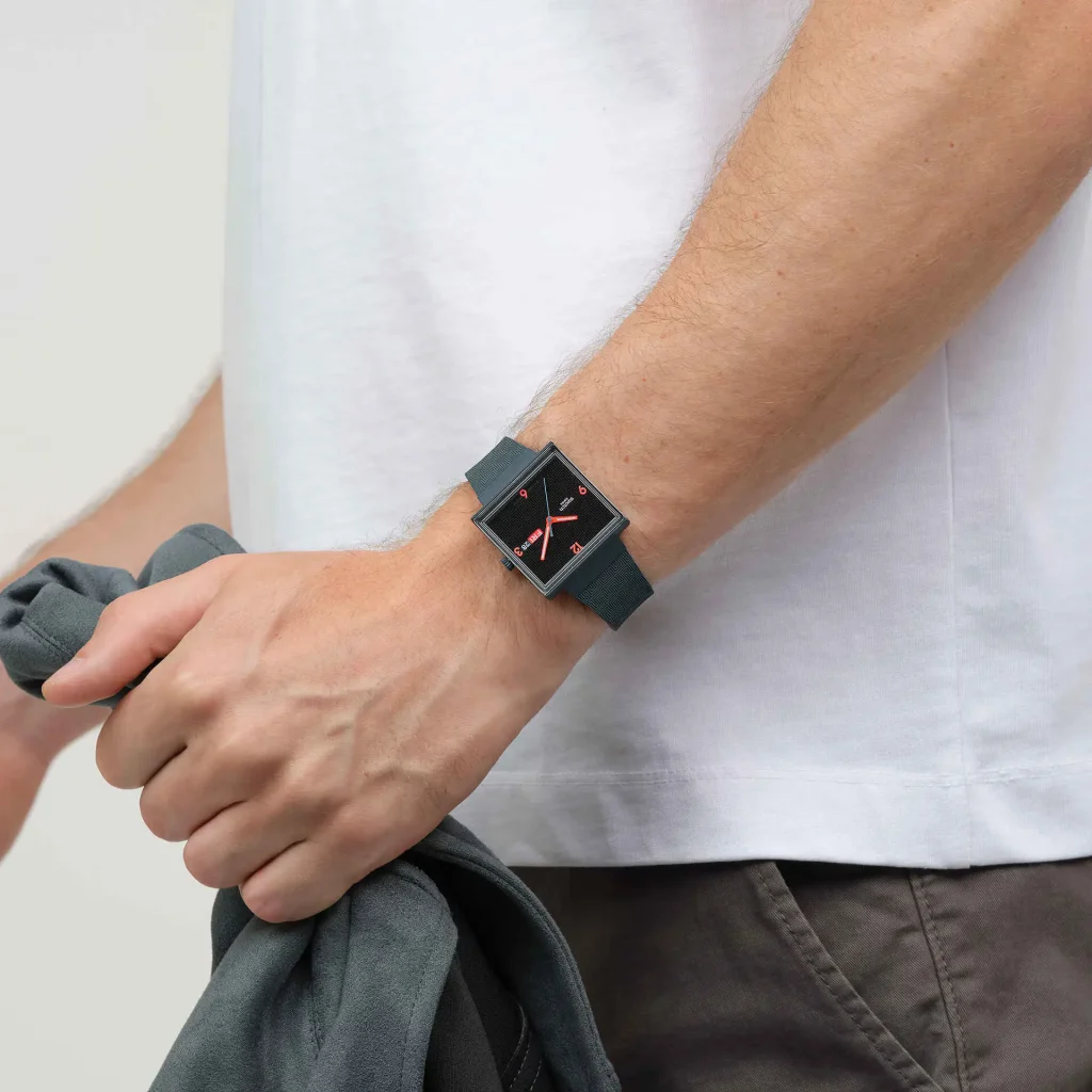 Homem com relógio quadrado e camisola cinzenta na mão.