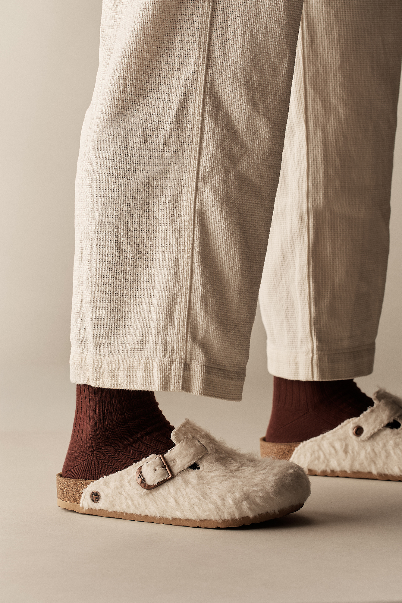 Pés calçados com os socos Birkenstock Boston Cole Bennett em feltro off-white com fivela de cobre, meias caneladas bordô e calças beges texturizadas