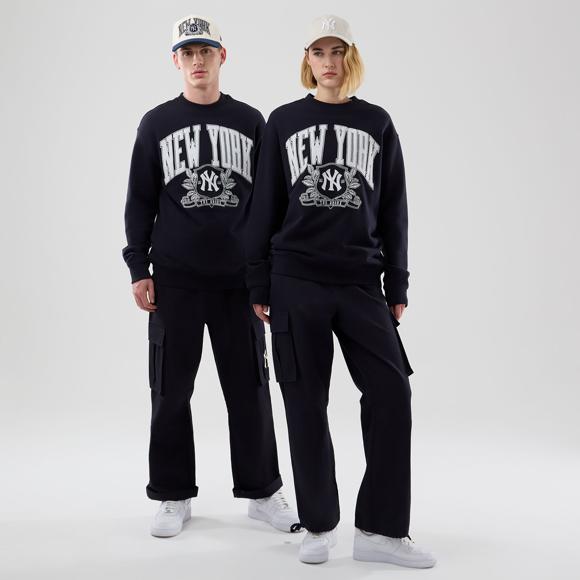 Homem e mulher a vestir sweatshirts oversized navy da coleção outono New Era com o logótipo New York Yankees, calças cargo escuras e bonés 9FORTY claros