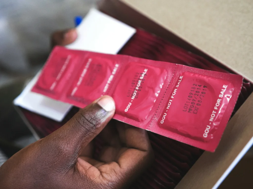 Mãos seguram uma tira de embalagens vermelhas de preservativos sobre uma caixa de cartão para escolher a largura nominal.