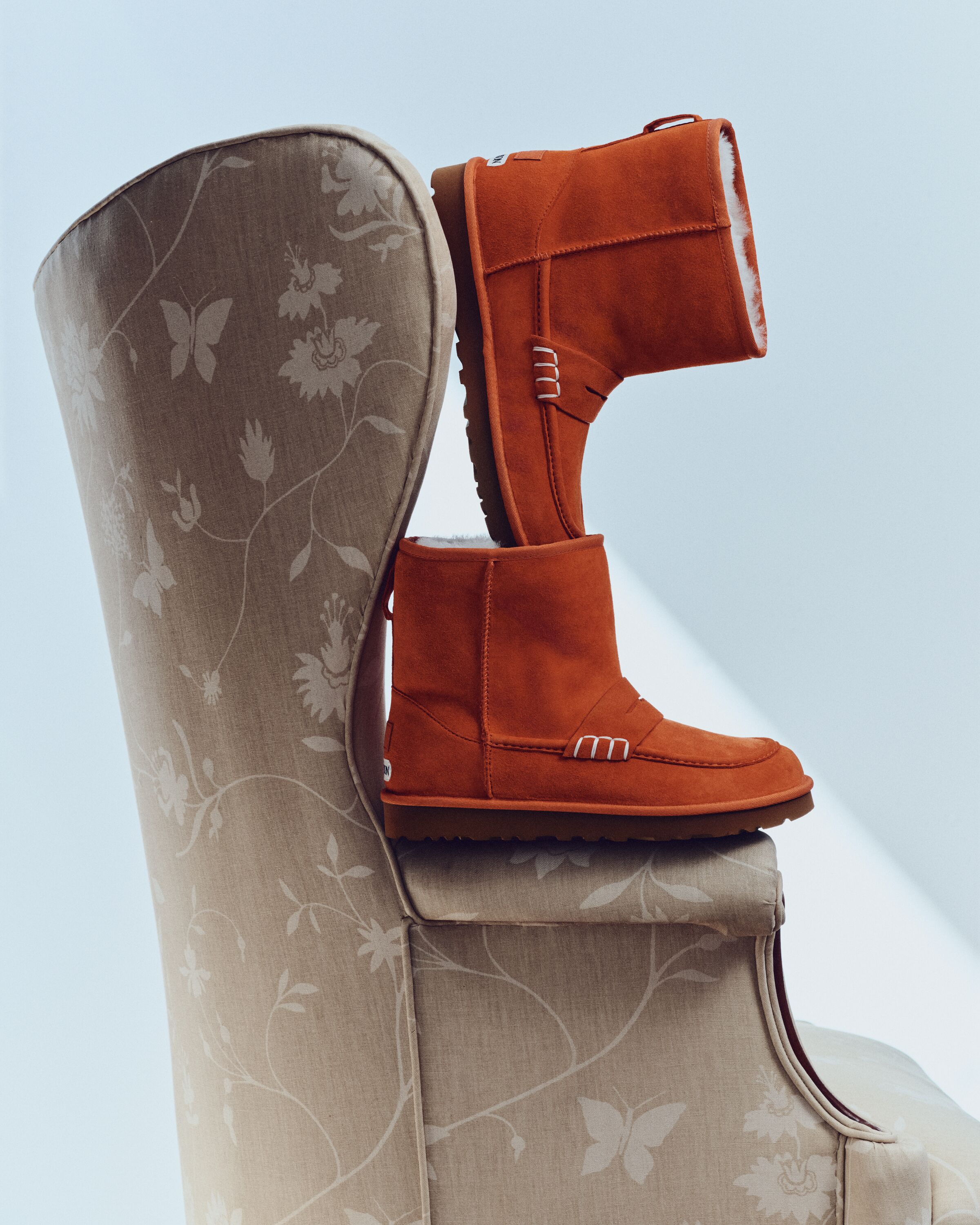 Botas UGG JW Anderson Loafer Boot em camurça laranja com interior em pelo empilhadas numa poltrona bege com padrão floral