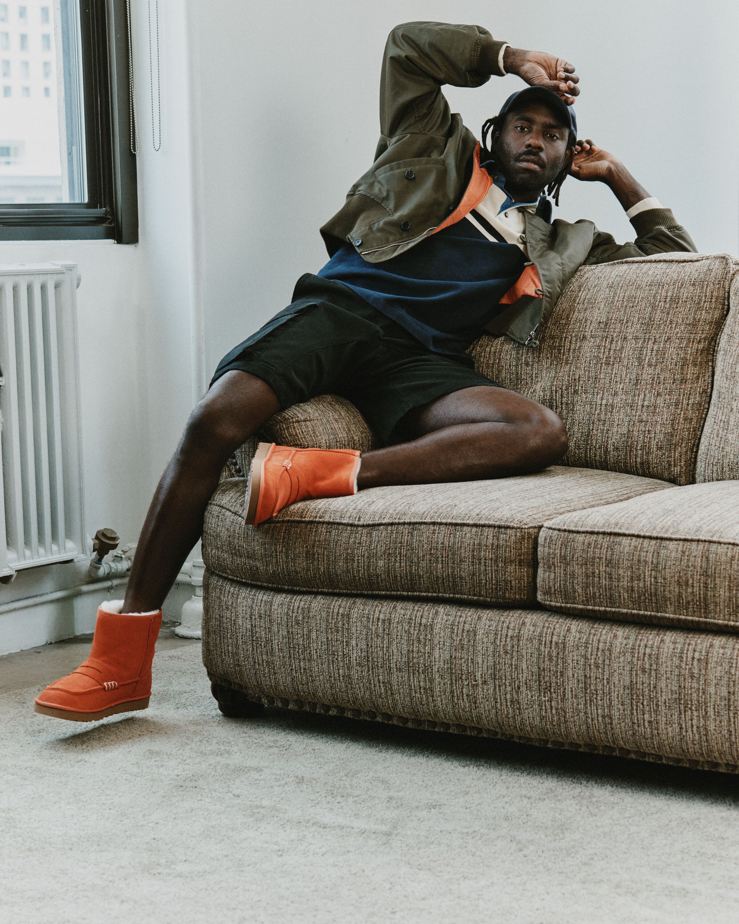 Homem sentado num sofá a vestir casaco verde, calções pretos e botas laranja UGG JW Anderson Loafer Boot.
