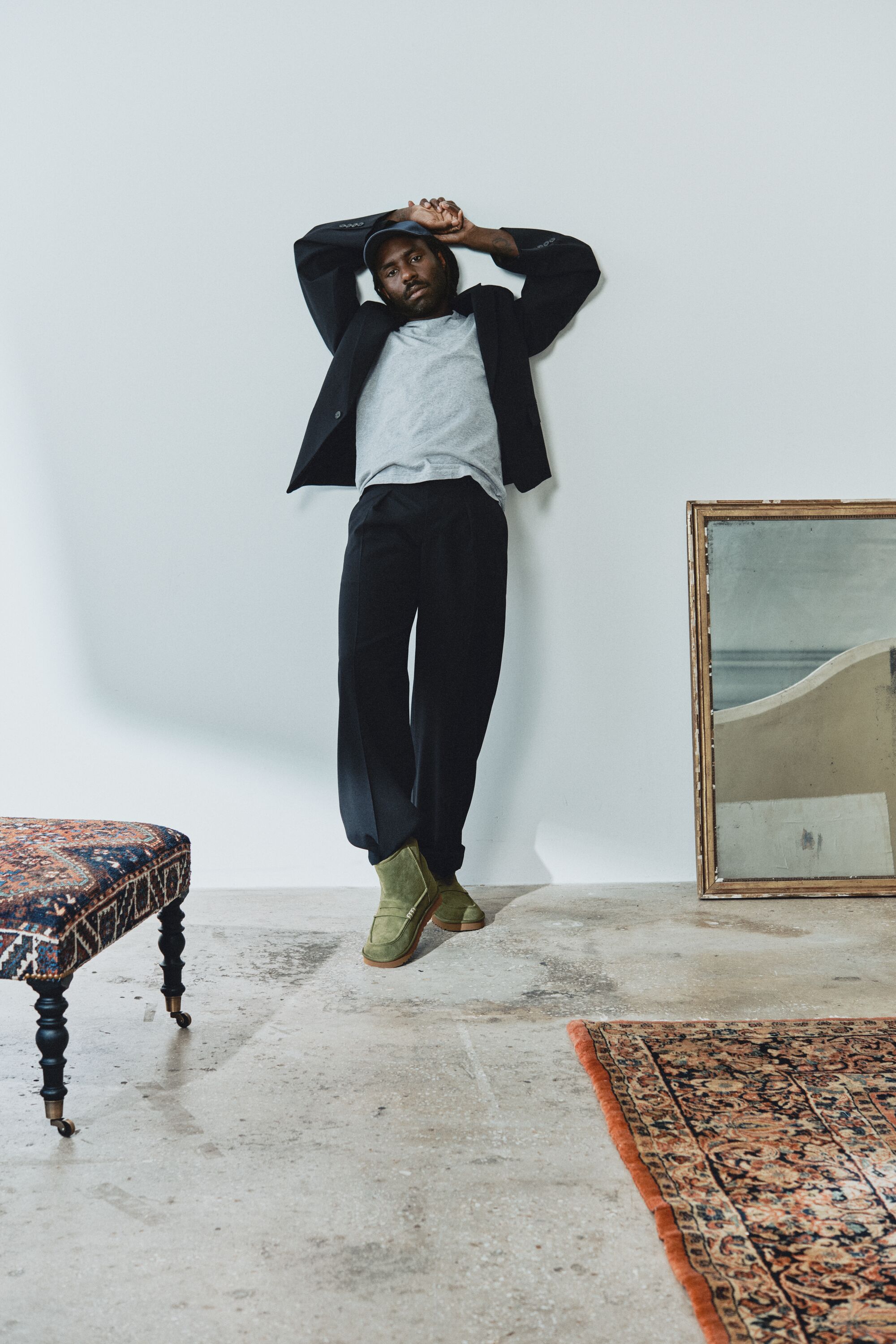 Homem encostado a uma parede branca a usar as botas verdes UGG JW Anderson Loafer Boot com calças escuras, t-shirt cinzenta e casaco.