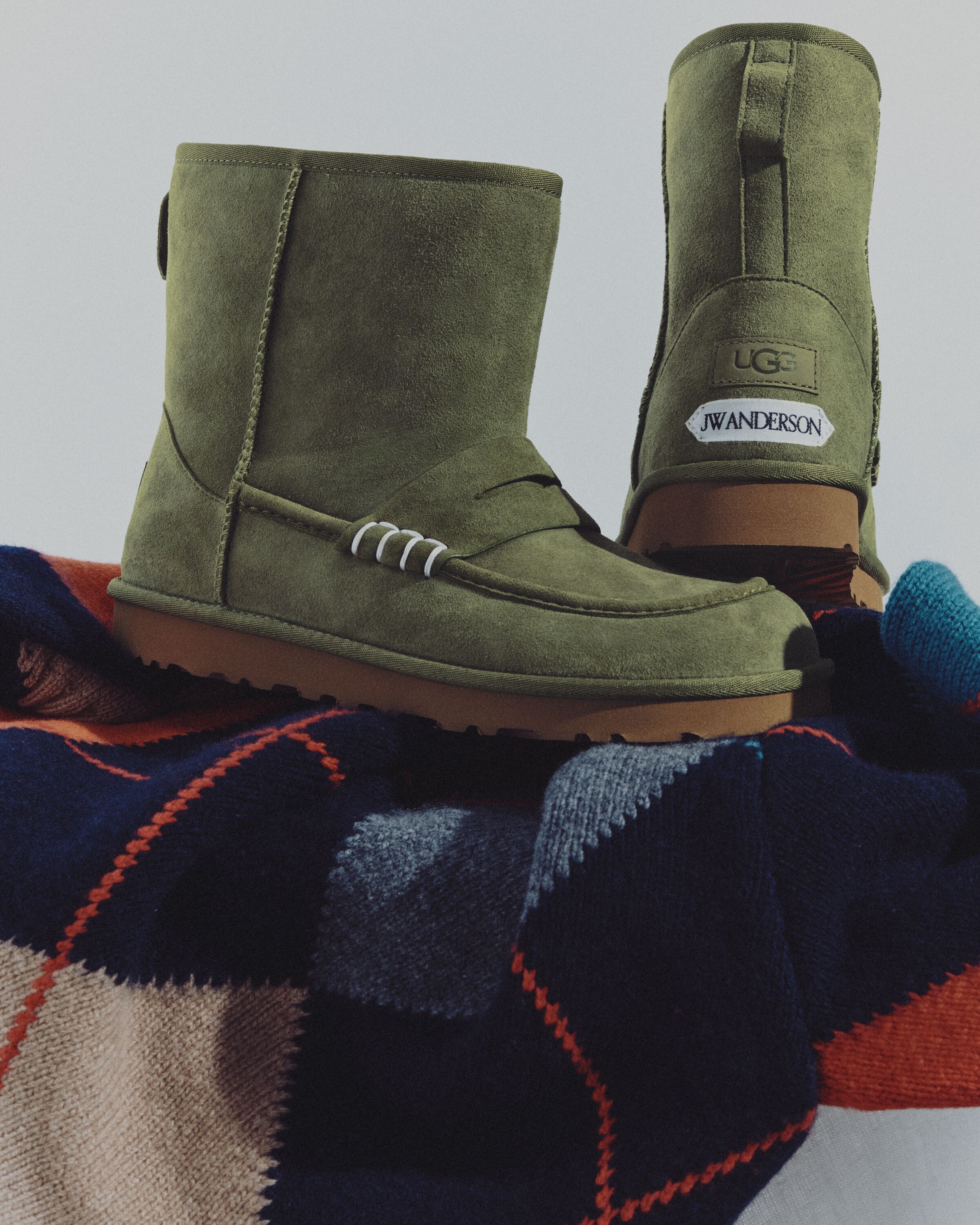 Par de botas UGG JW Anderson Loafer Boot em camurça verde com costuras brancas e logótipos no calcanhar, sobre uma manta de malha com padrão.