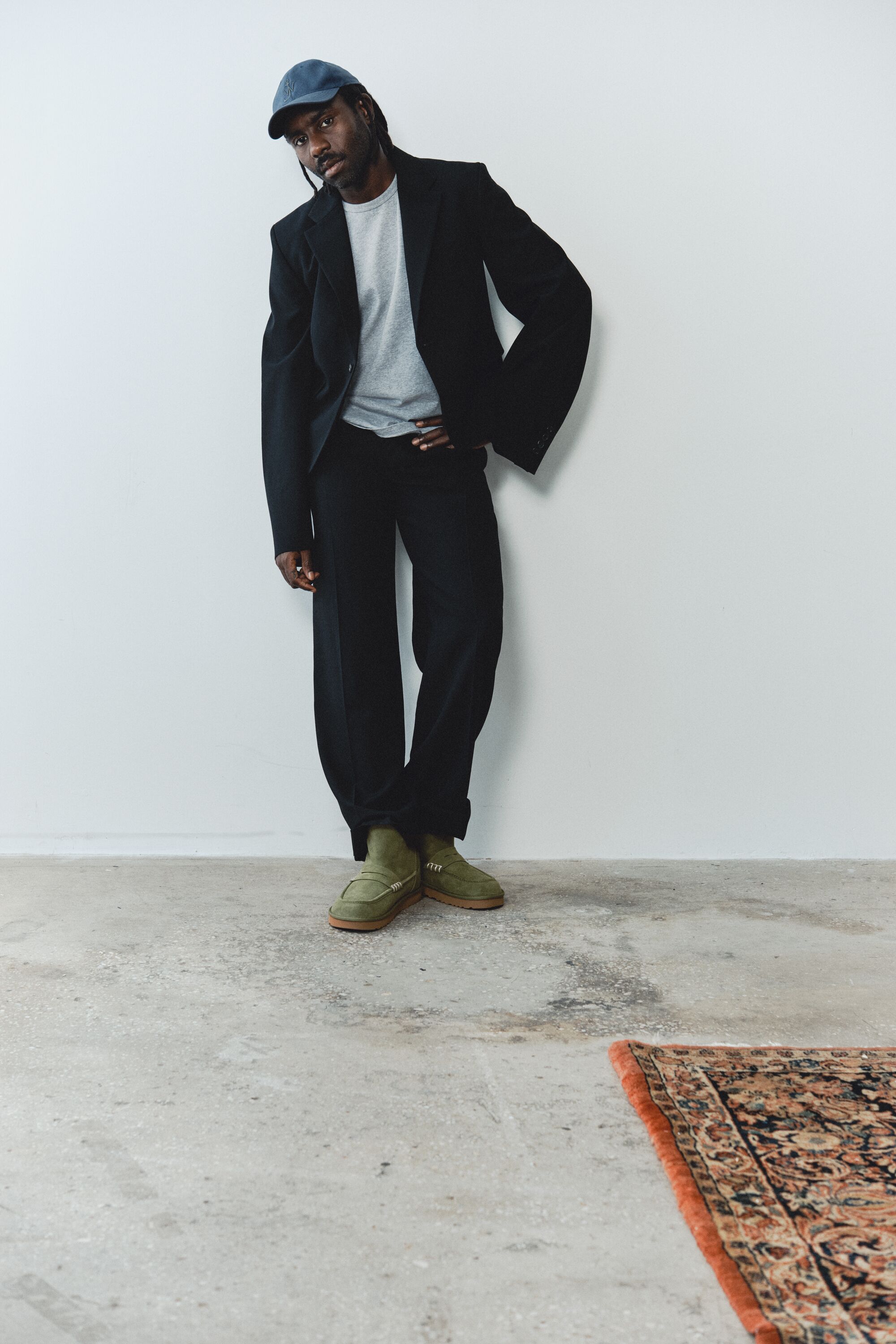 Homem com fato preto e boné azul a calçar as botas verdes UGG JW Anderson Loafer encostado a uma parede branca