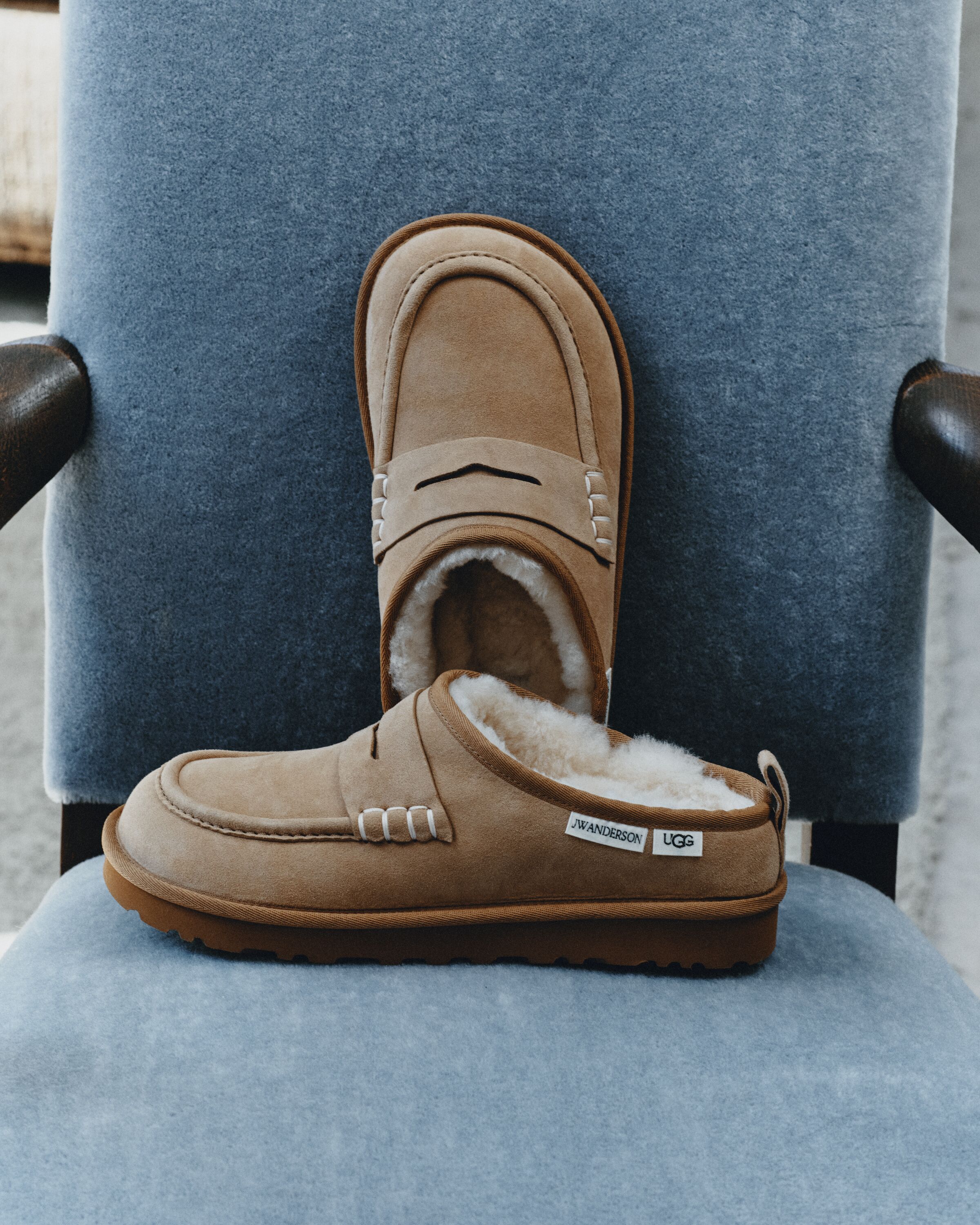 Par de sapatos UGG JW Anderson Loafer em camurça castanha com interior em pelo e sola de borracha pousados numa cadeira de veludo azul.