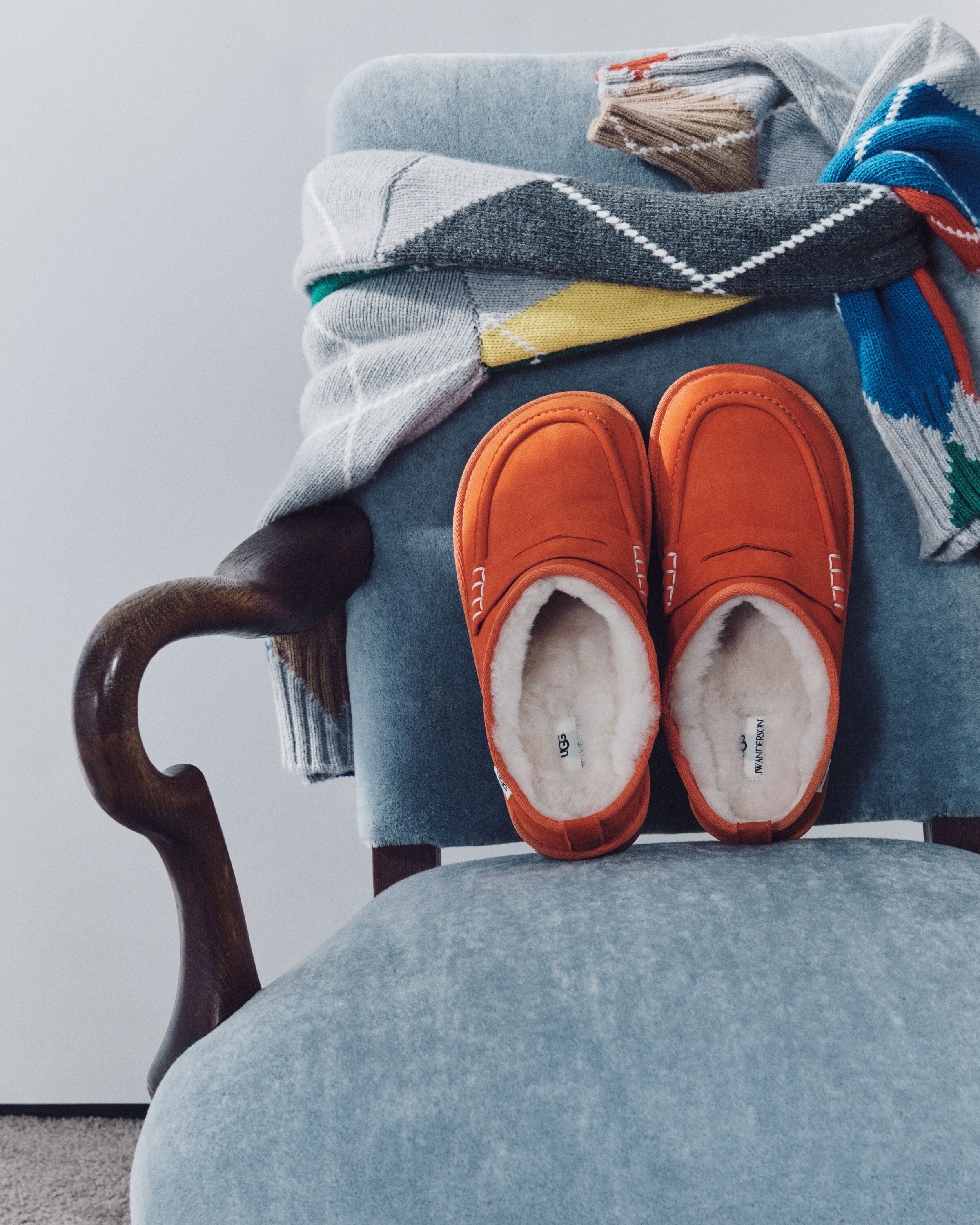 Par de sapatos UGG JW Anderson Loafer em camurça laranja com interior em pelo branco e costuras brancas, pousados numa cadeira de veludo azul claro com uma camisola de malha estampada nas costas.