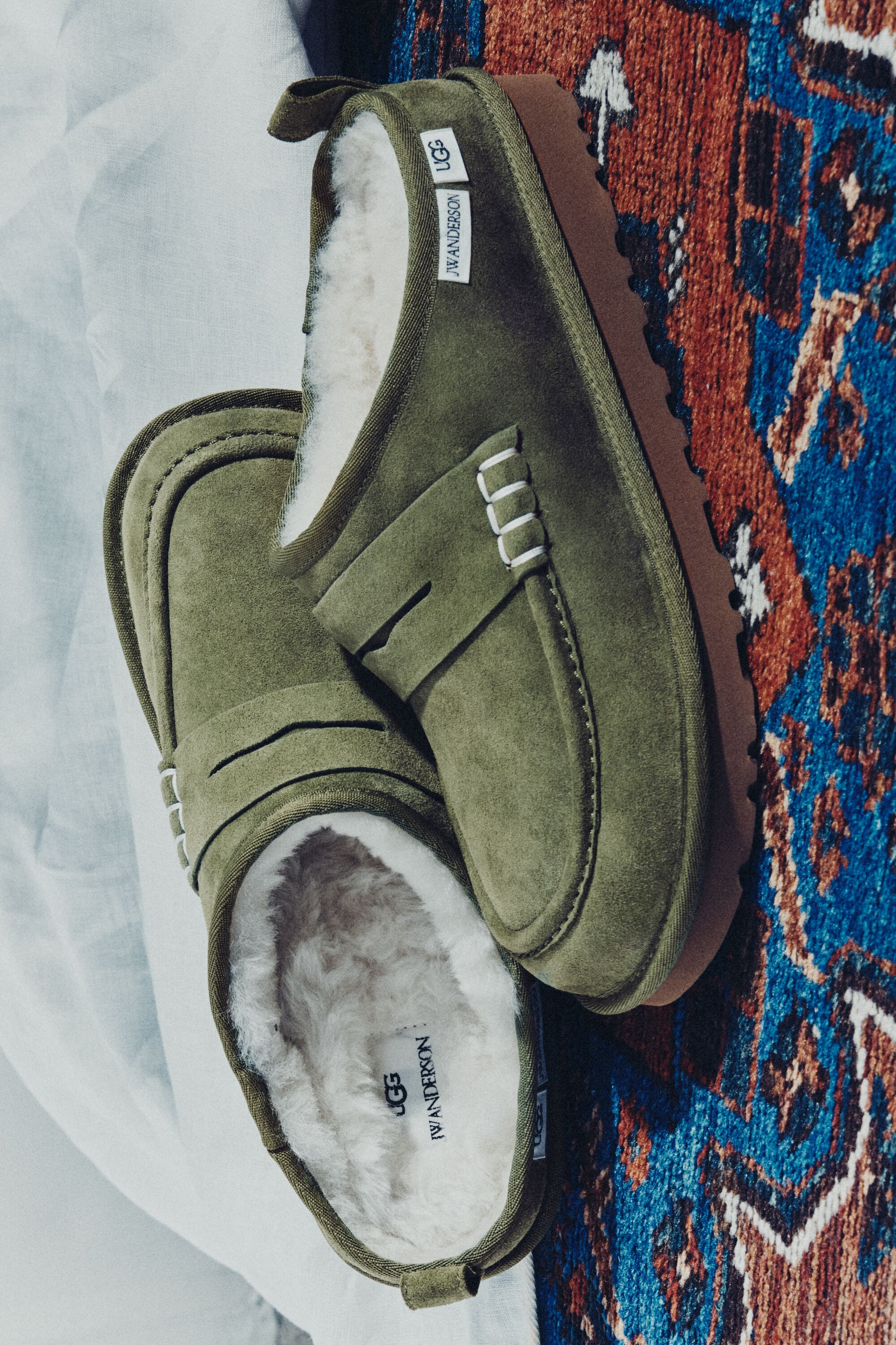 Par de sapatos UGG JW Anderson Loafer em camurça verde com interior em pelo branco sobre tapete estampado e tecido claro