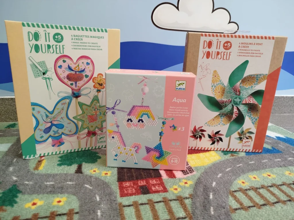 Três kits de trabalhos manuais Djeco sobre um tapete infantil.